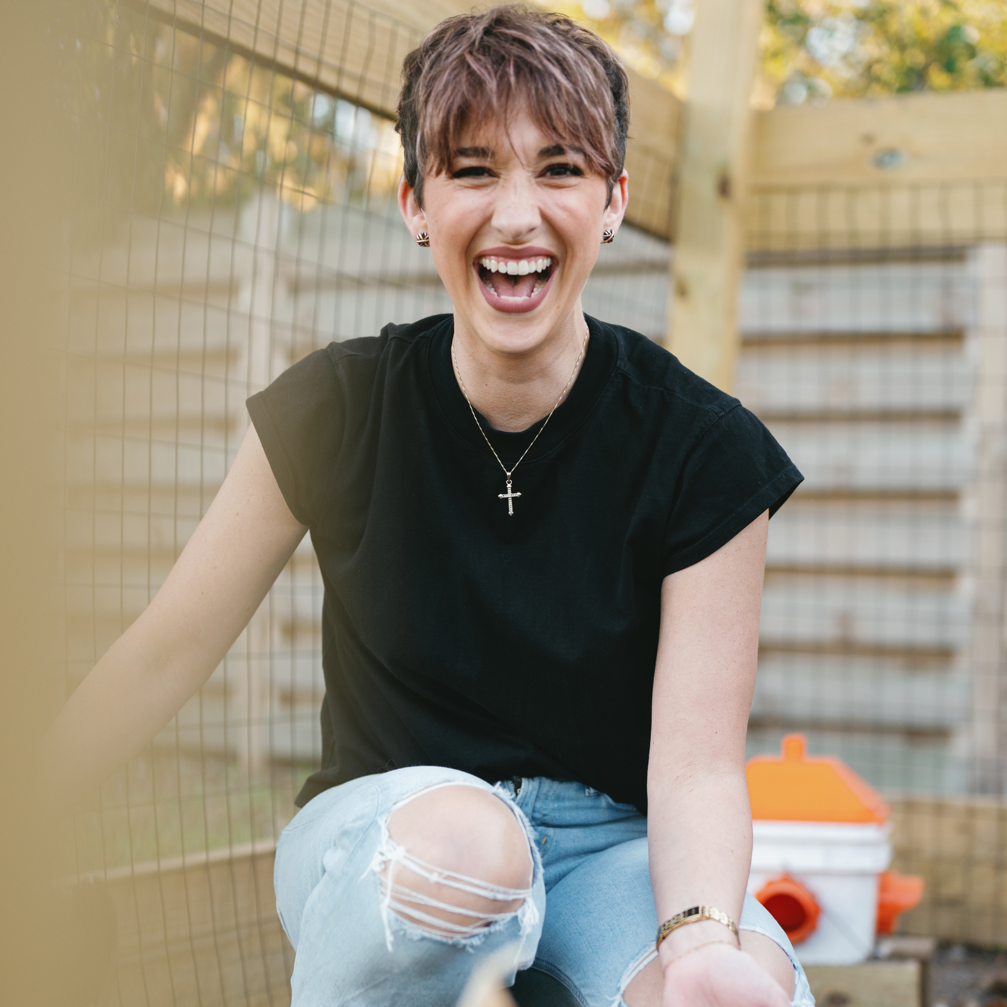 girl with pixie wearing black t shirt and ripped blue jeans laughing in the chicken coop