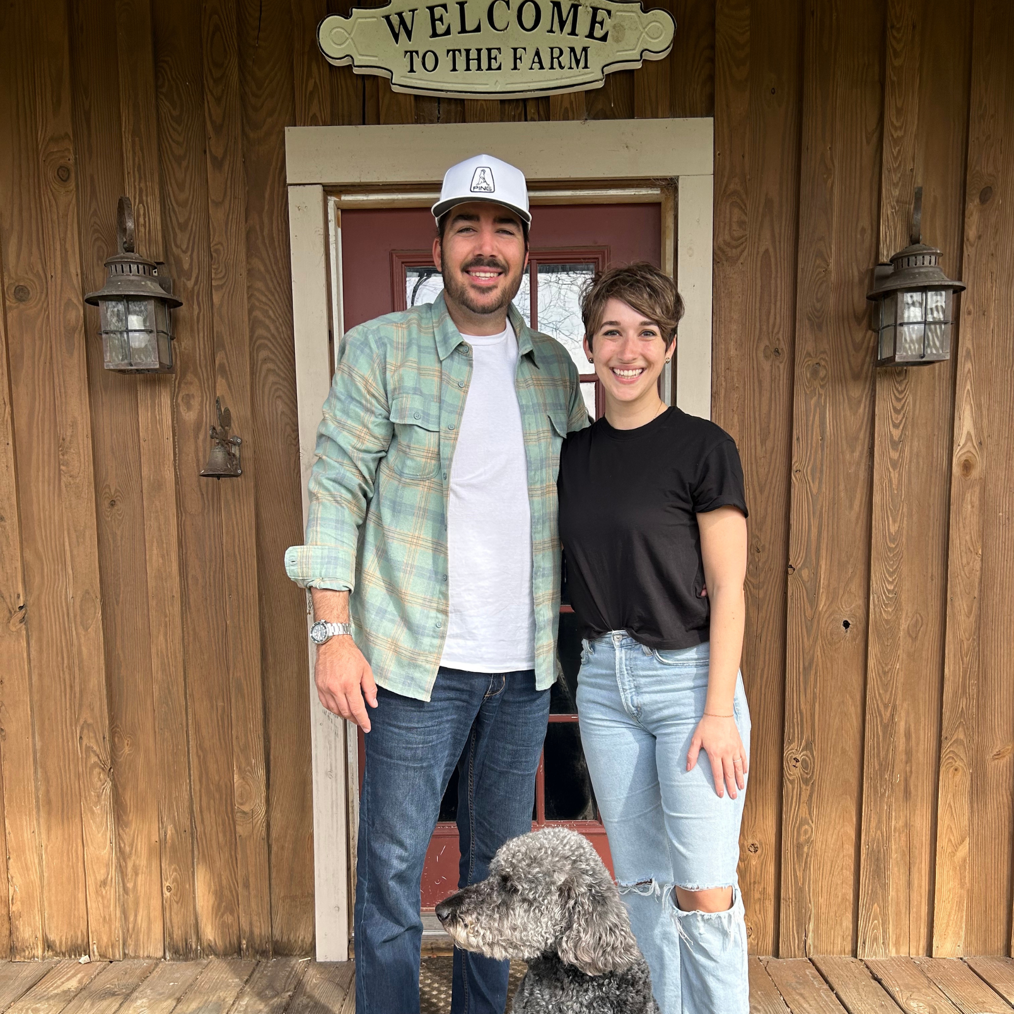 husband, wife and goldendoodle standing at front door of home with welcome to the farm sign